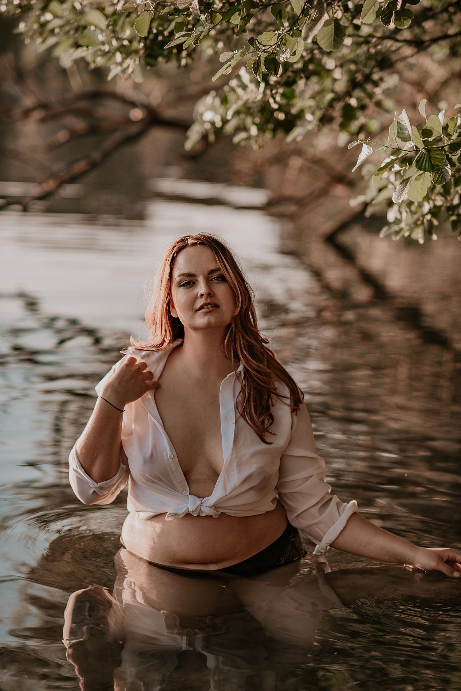 Woman in white shirt posing in water with tree branches overhead in artistic boudoir photo.