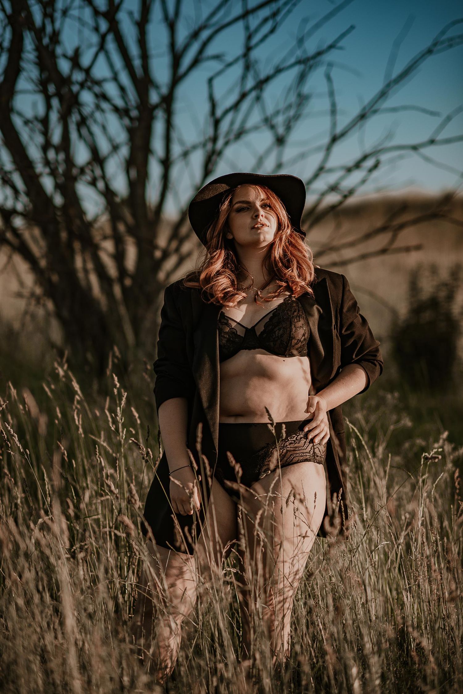 A moody fashion portrait in tall grass with a dark brimmed hat against a sunset sky.