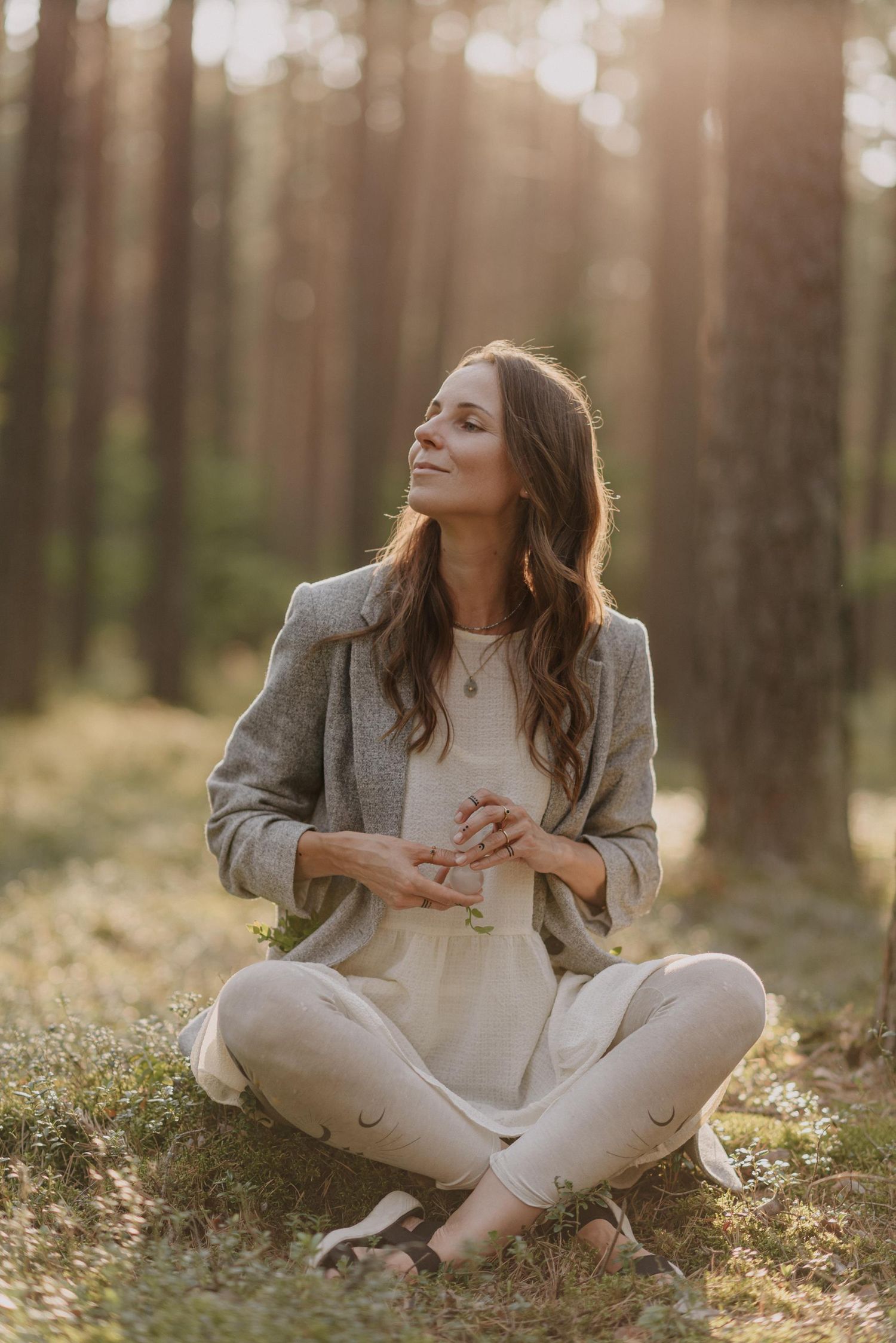 Person in white dress and gray blazer sitting cross-legged in a sunlit forest.