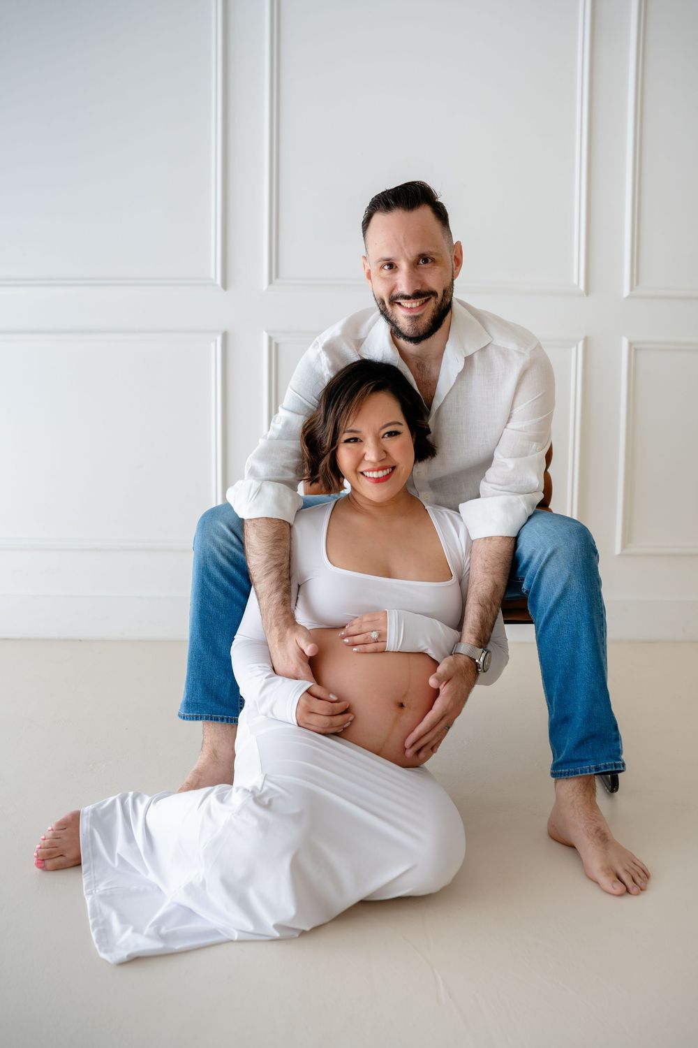 Minimalist maternity photos showing expectant couple in casual white attire sitting on floor.