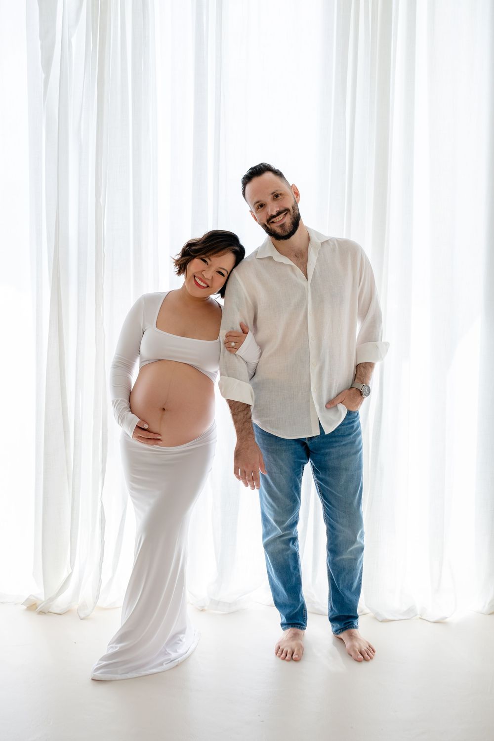 Artistic maternity photo series showing couple in white against bright curtain backdrop.
