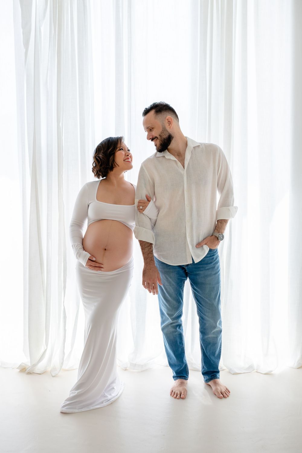 A couple shares an intimate moment during a maternity photoshoot against white curtains in a bright studio setting.