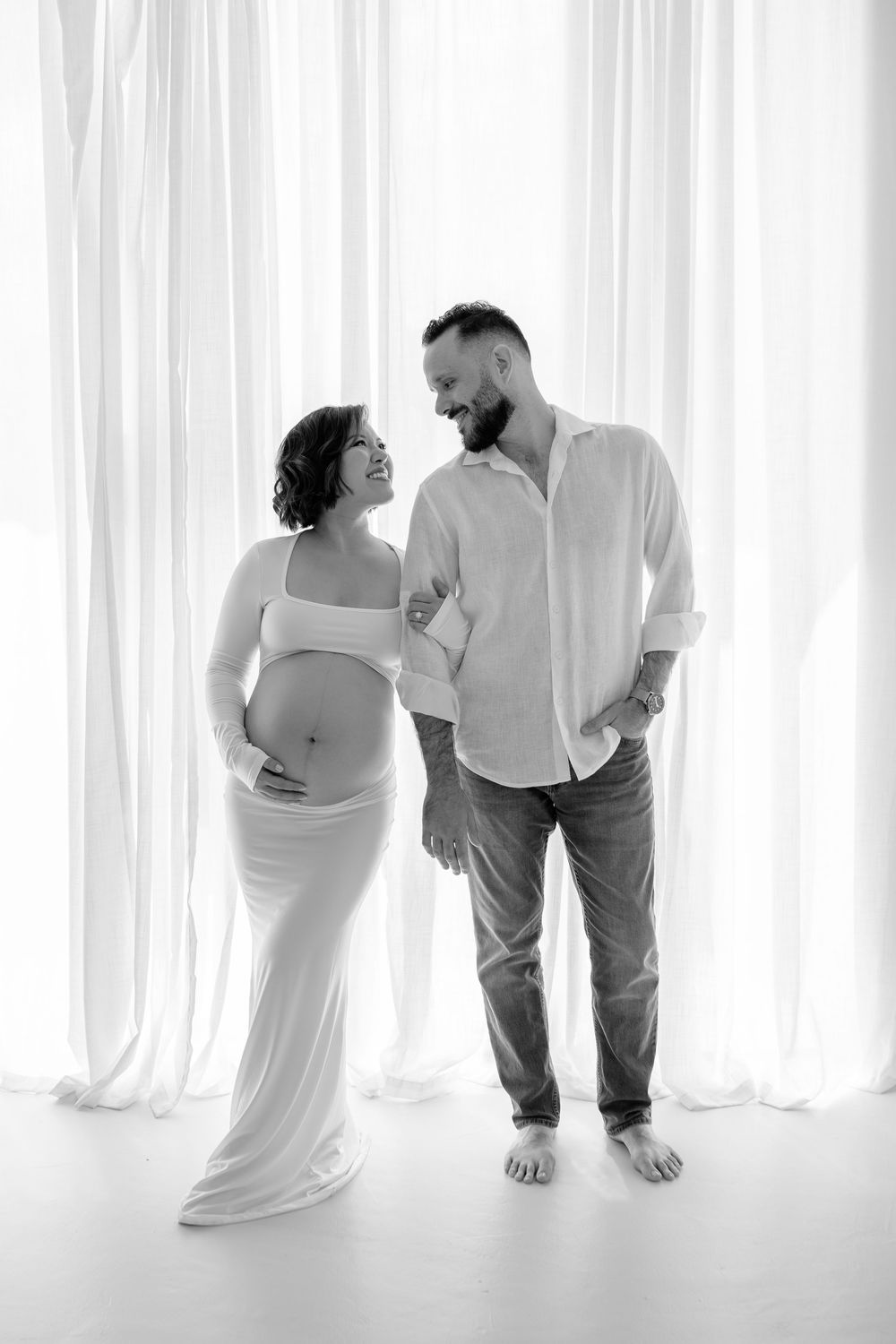 Black and white maternity portraits with couple posing playfully against sheer white curtains.