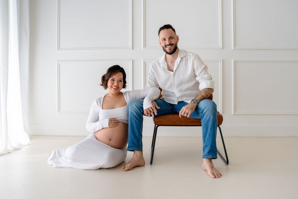 Expecting couple in casual attire poses together in bright white studio setting with wainscoting.