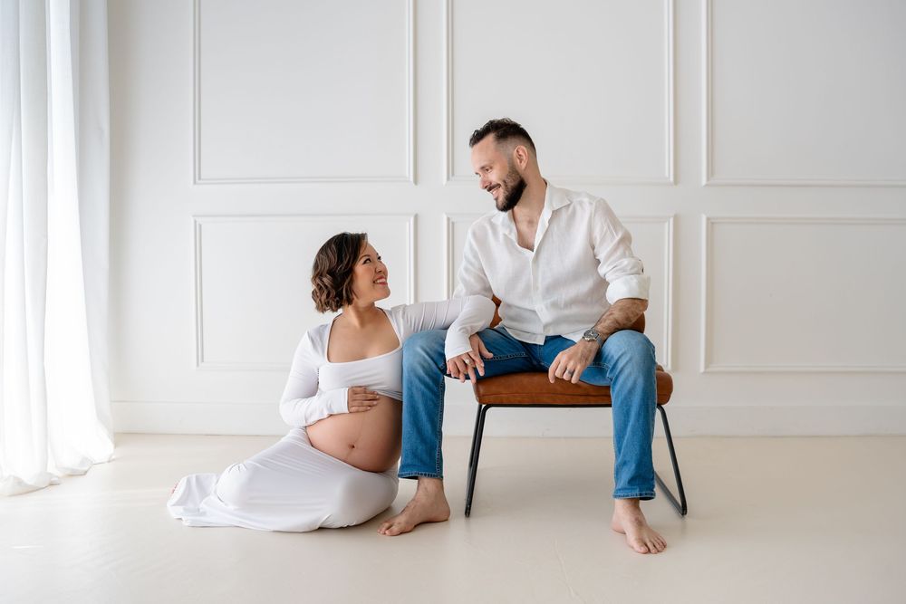 Minimalist white studio maternity photo shoot with couple sitting on floor against paneled wall.