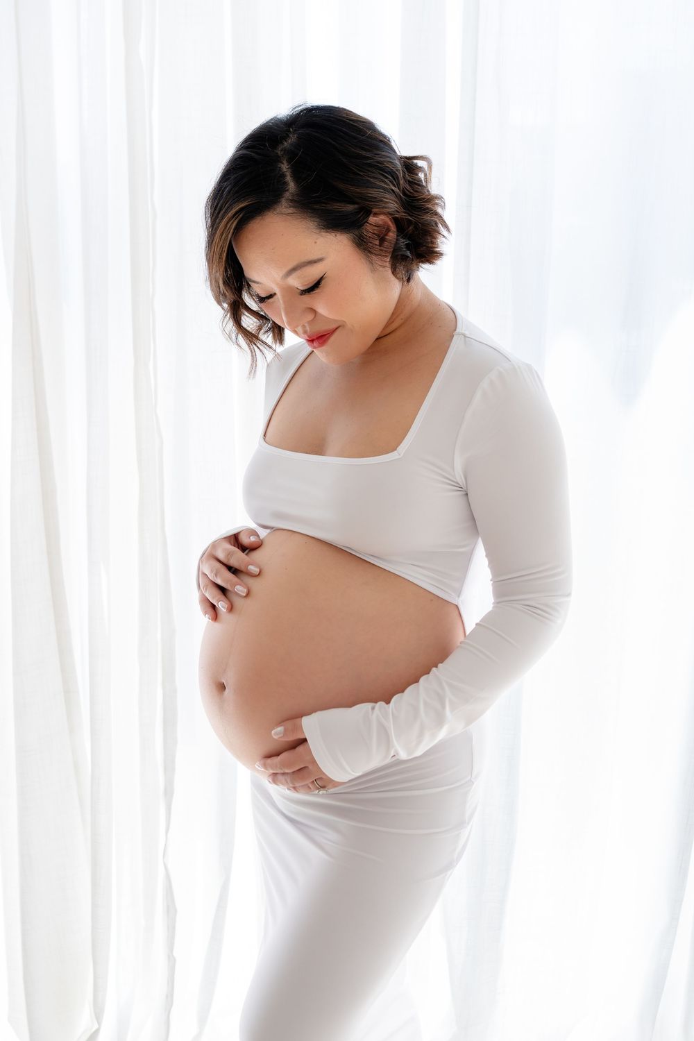 Artistic black and white maternity photos showing pregnant woman in white top cradling her belly against sheer curtains.