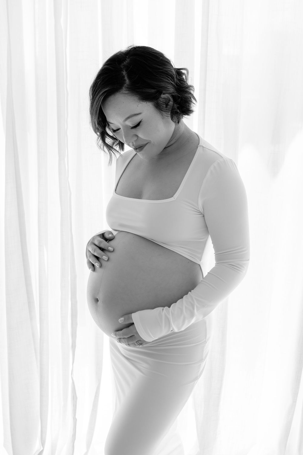Artistic maternity photoshoot featuring an expectant mother in white against a bright window backdrop.