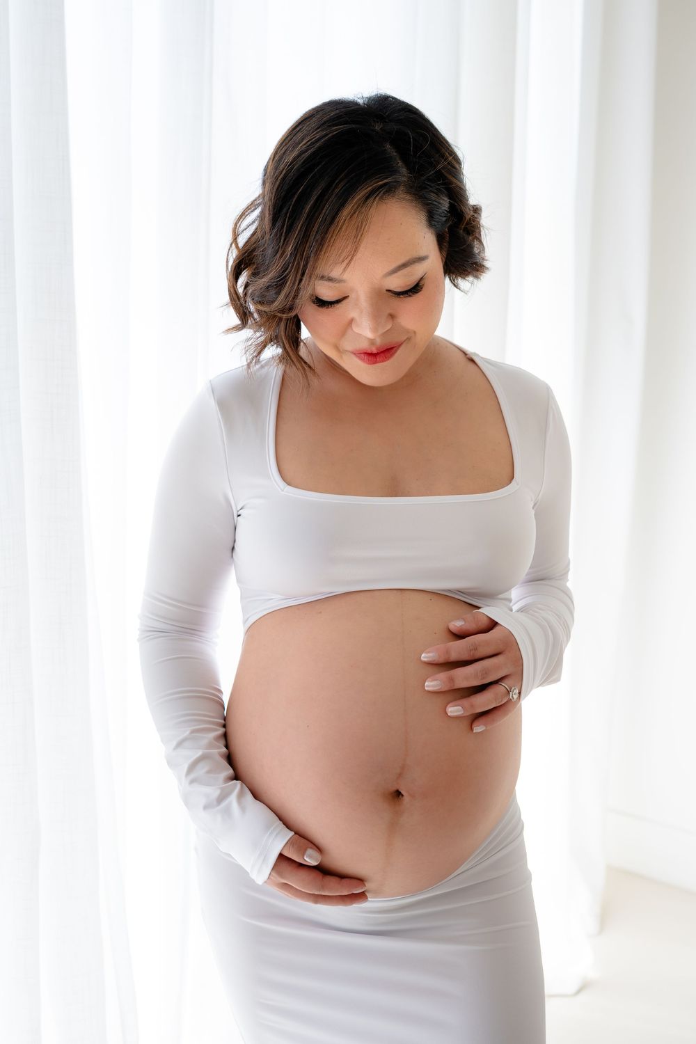 Intimate maternity portrait showing a pregnant woman in white clothing touching her belly near sheer curtains.