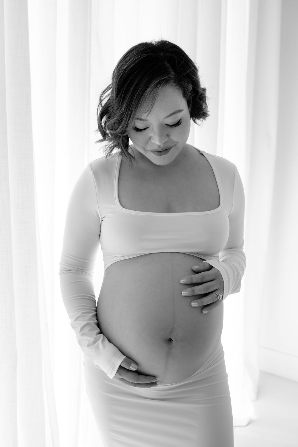 Black and white maternity photo of an expectant mother in a white dress looking down at her belly with a gentle smile.