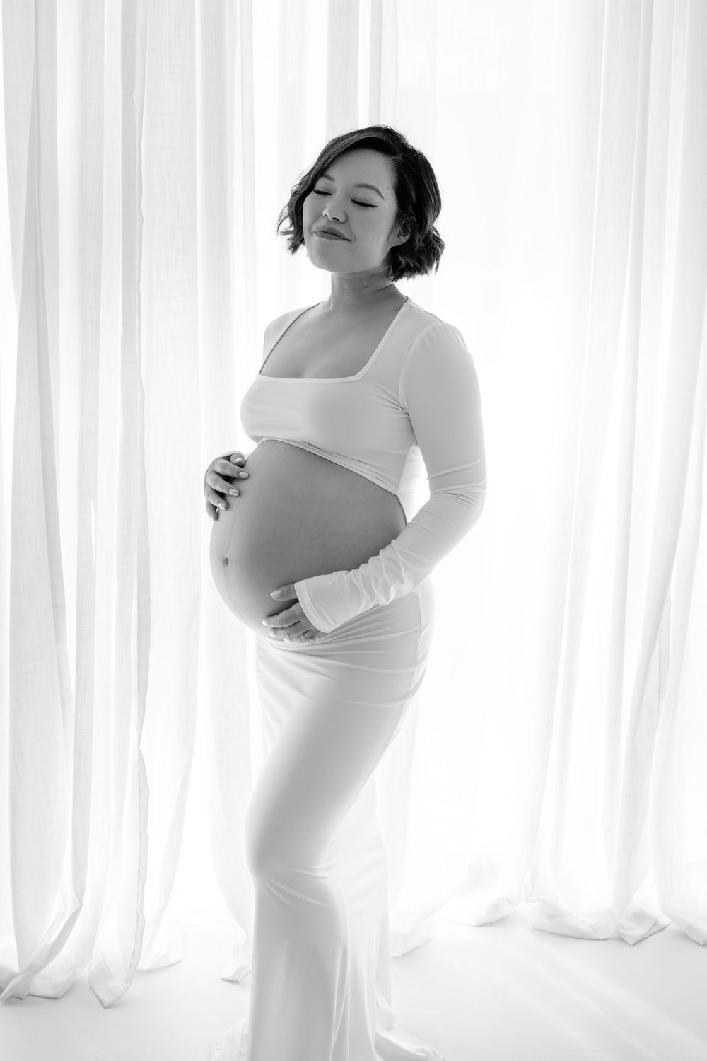 Elegant black and white maternity photo series with soft window lighting and flowing white fabric.