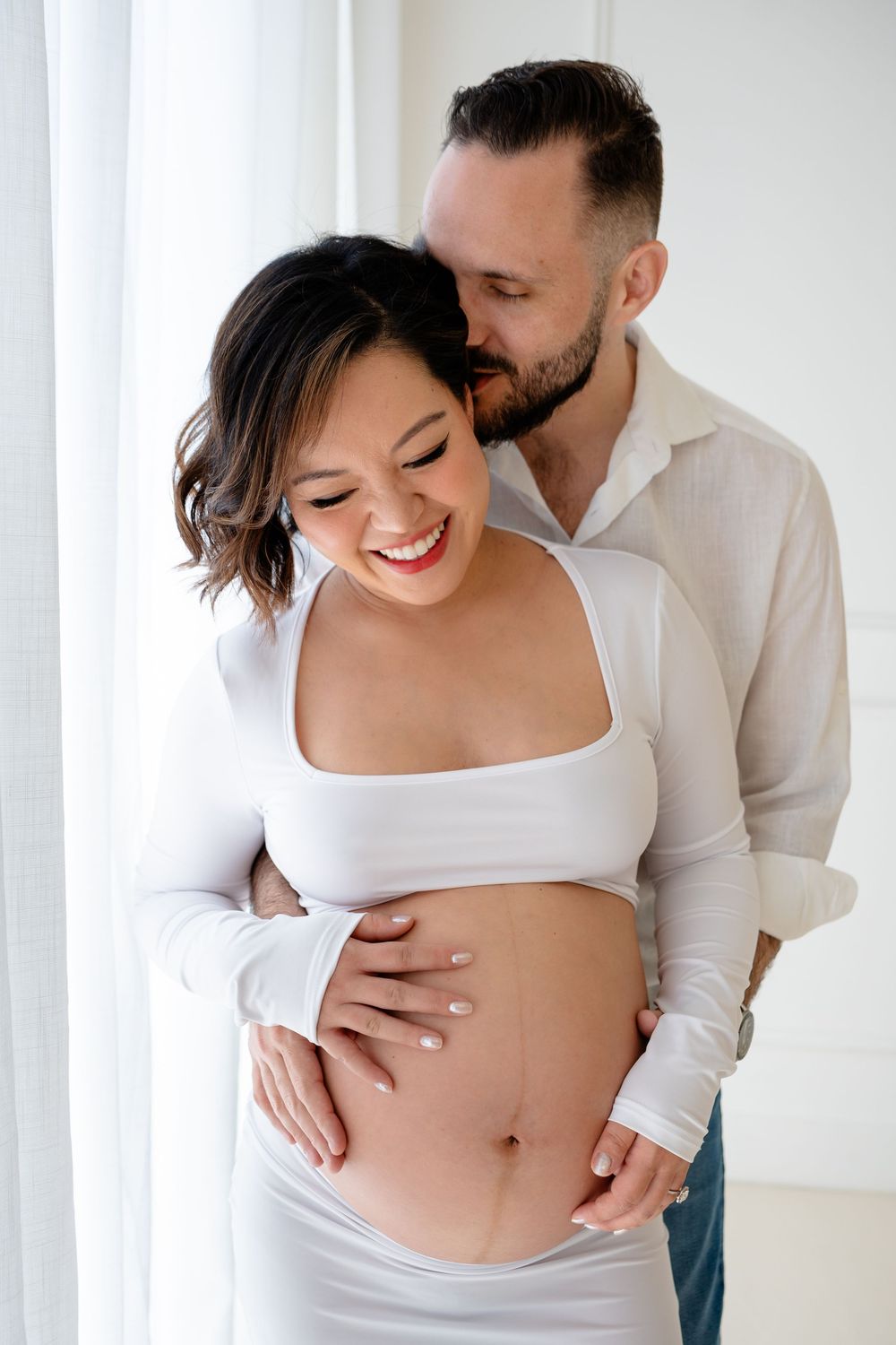Intimate maternity photoshoot showing couple in white clothing embracing against bright interior wall.