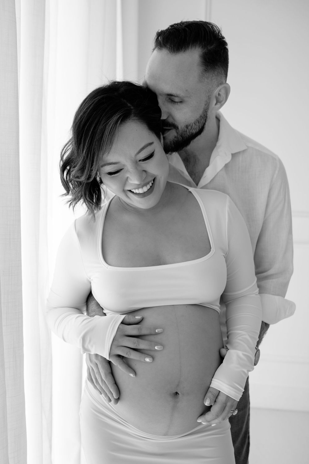 Intimate black and white maternity photos of couple embracing with woman wearing white top against light background.