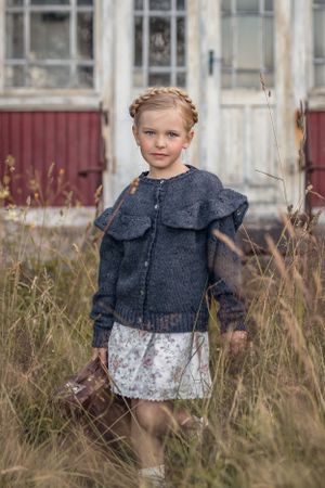 Vintagefoto. En flicka i gammeldags kläder står framför ett gammalt torp.