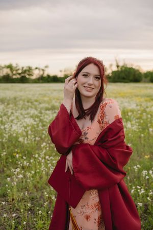 Young woman with red hair in floral dress and red shawl smiling in a blooming meadow at sunset.