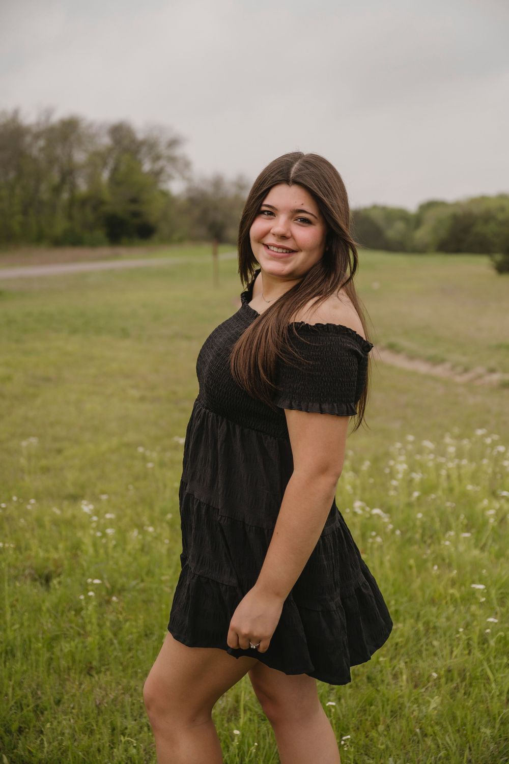 Young woman in black off-shoulder mini dress smiling in a green field with wildflowers on a cloudy day.