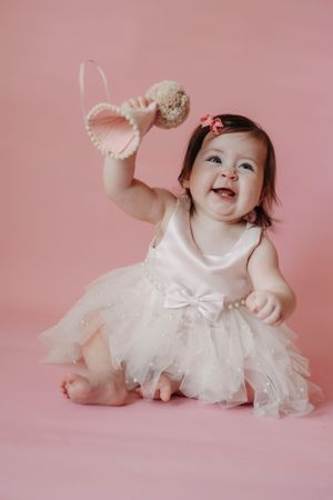 Baby in white tulle dress laughs and plays on pink background during photo session.