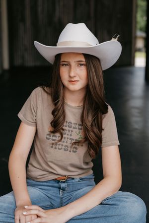 Girl in cowboy hat and t-shirt sits casually in a rustic setting.