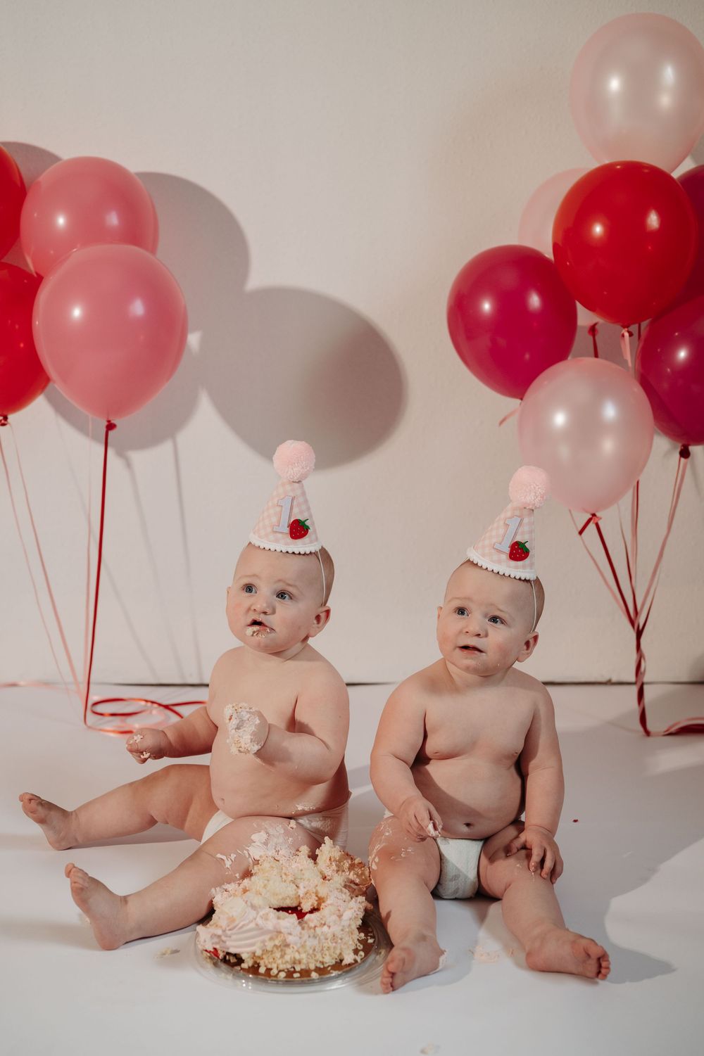 Twin babies having cake smash photo session with pink and red balloons in studio setting.