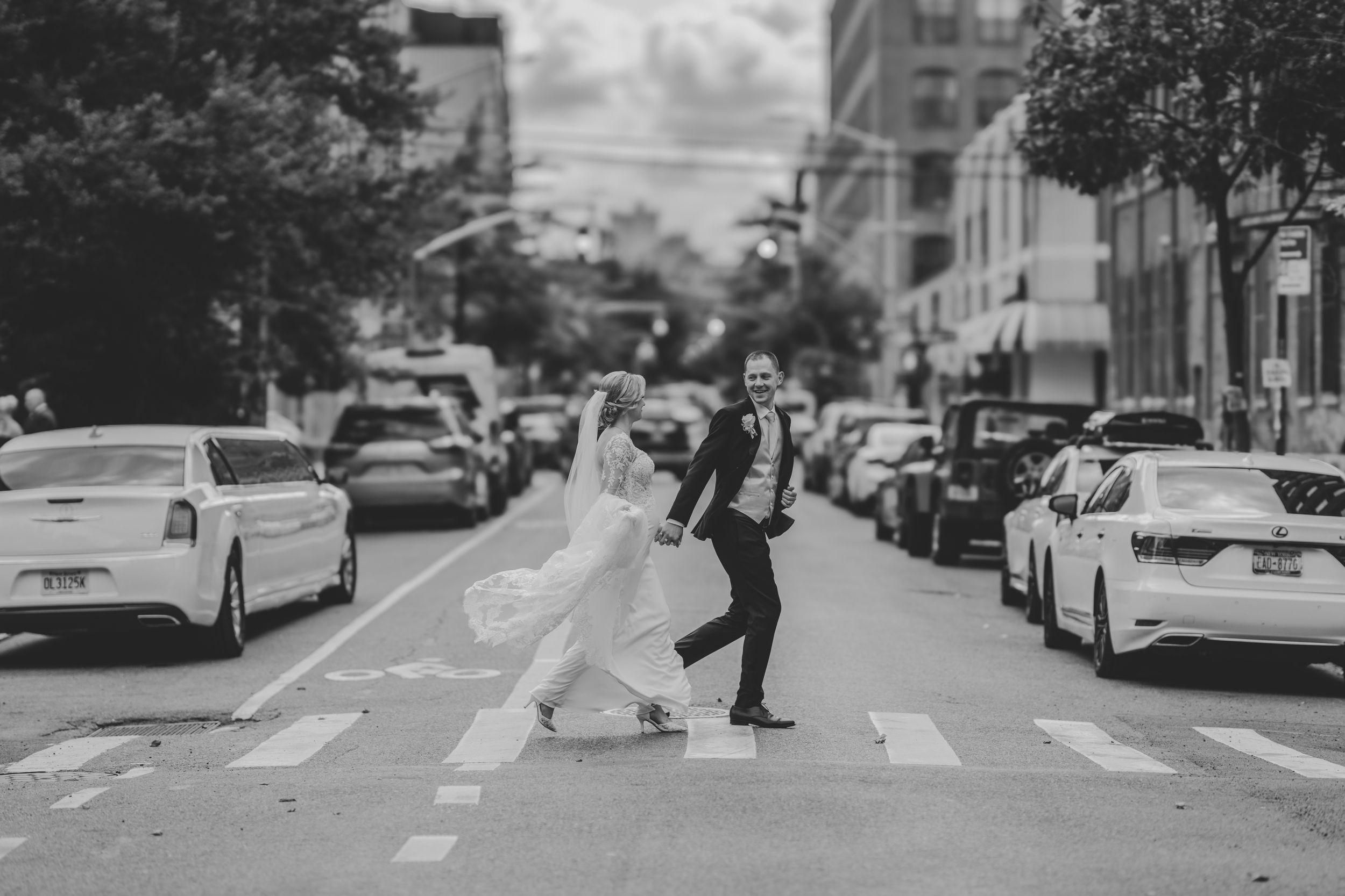 A couple in wedding attire crosses a city street with parked cars lining both sides in black and white.
