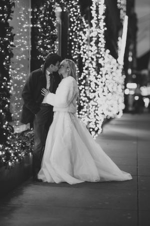 A romantic black and white wedding photo showing a couple embracing near cascading string lights in an evening setting.