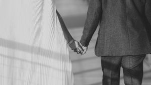 Close-up black and white image of two people holding hands, one wearing a white dress and one in a gray suit.