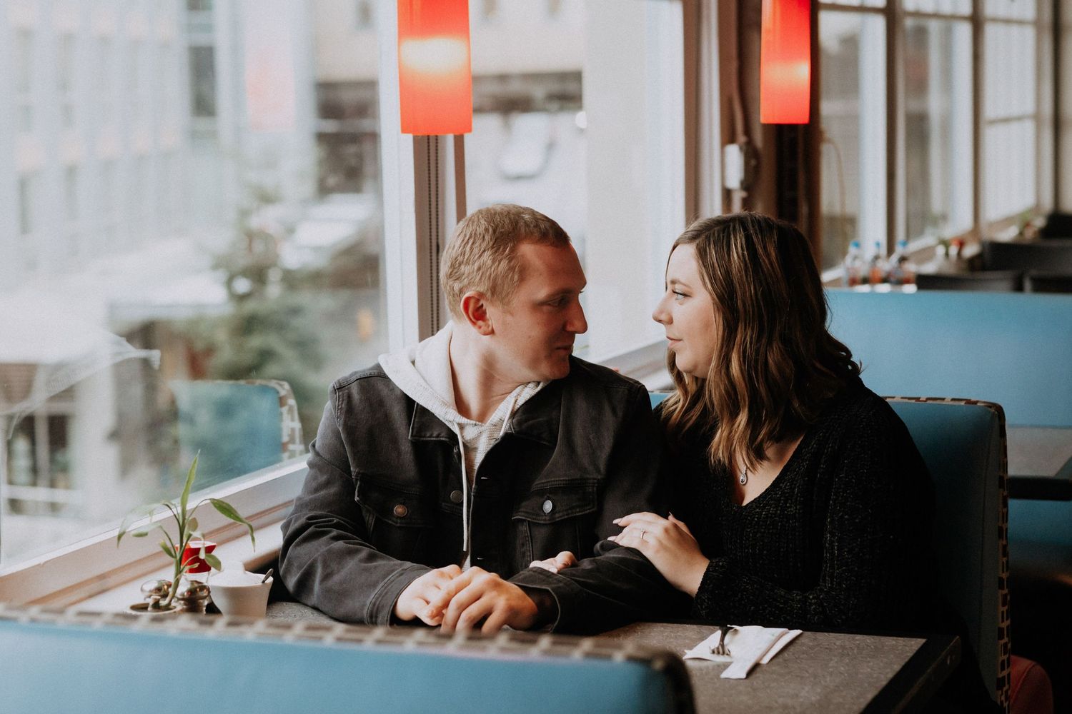 A couple sits together at a cafe table by a window, sharing an intimate conversation in warm lighting.