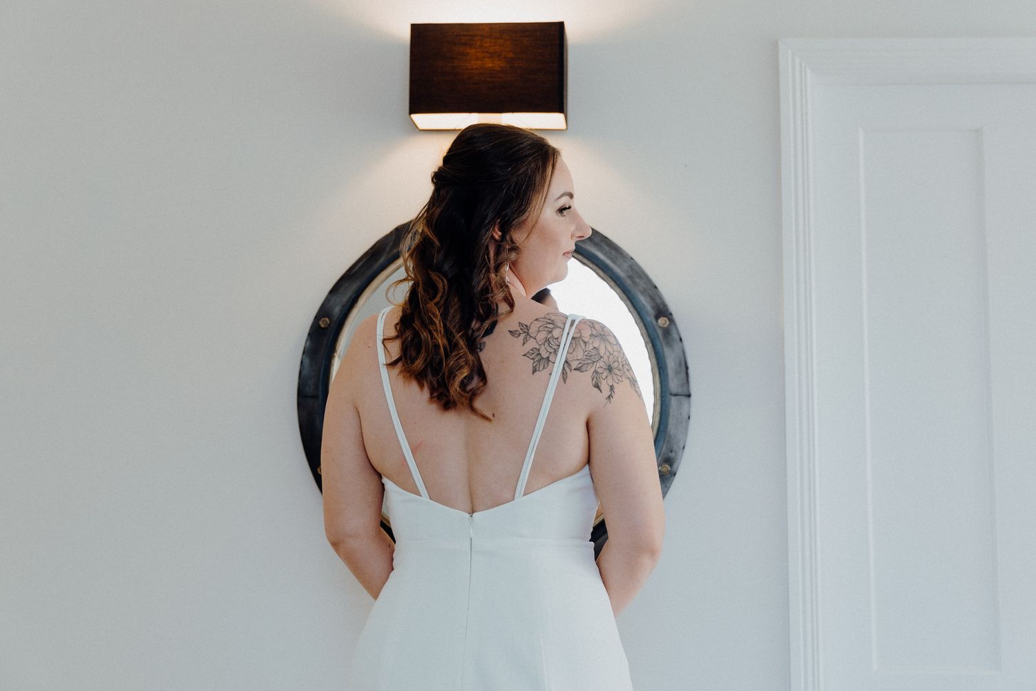 Back view of a bride in a white wedding dress standing next to a round mirror with a black wall lamp above, captured by Apertured Media.