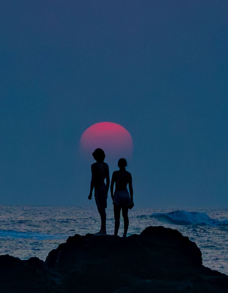 Two silhouettes stand on rock formation against large red sun and ocean horizon.
