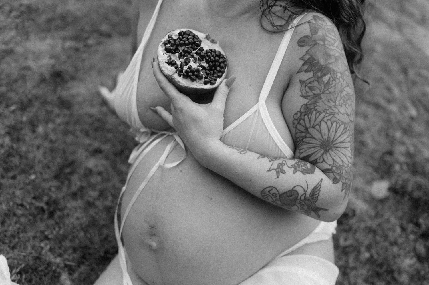 Pregnant woman in white dress holds a succulent plant in a black and white photo showing arm tattoos and baby bump.
