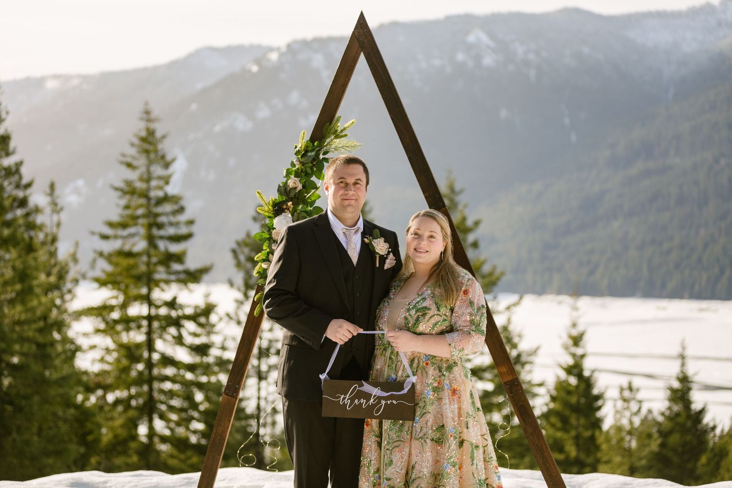 A couple poses in front of a triangular wooden arch with mountain and lake views in the background.
