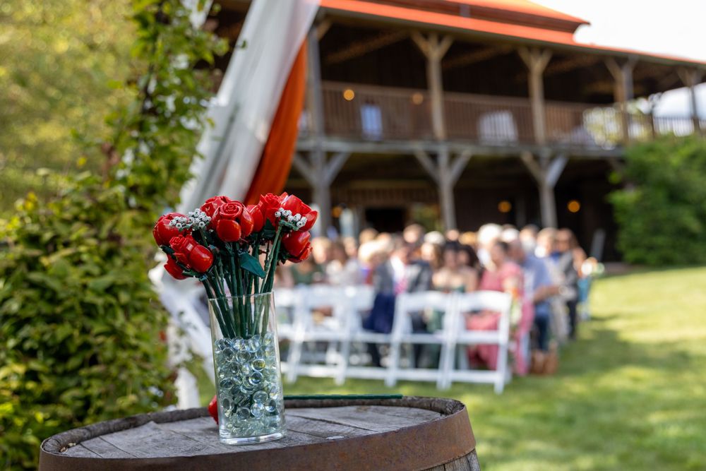 A vase of red carnations on a barrel with an outdoor wedding ceremony setup in the background under a covered pavilion.