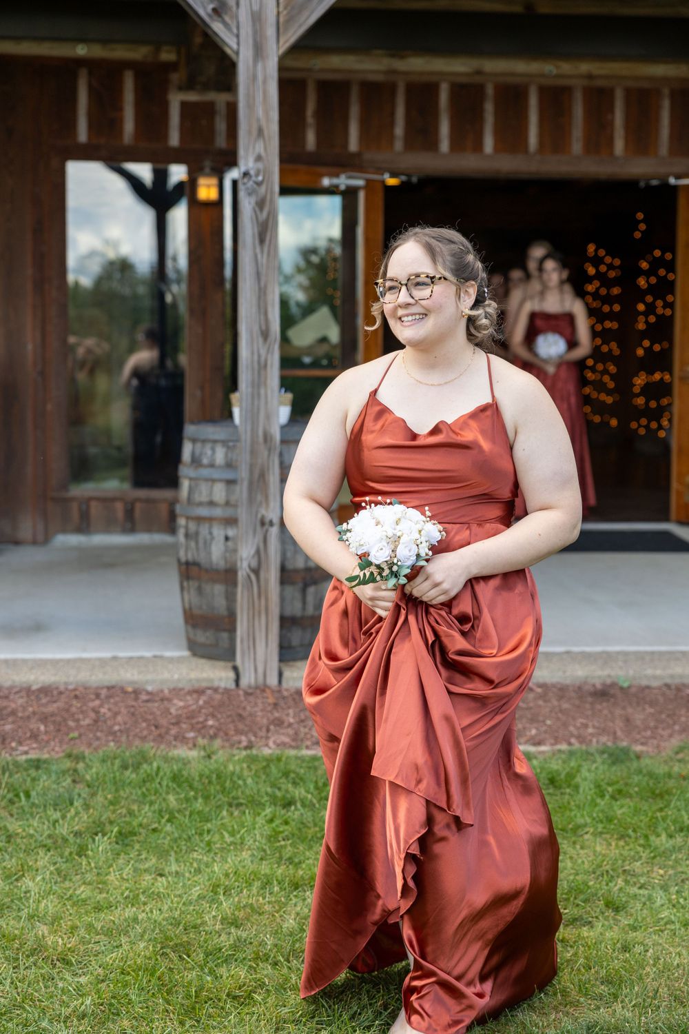 A bridesmaid in a flowing rust-colored dress holds a white bouquet outside a rustic venue.