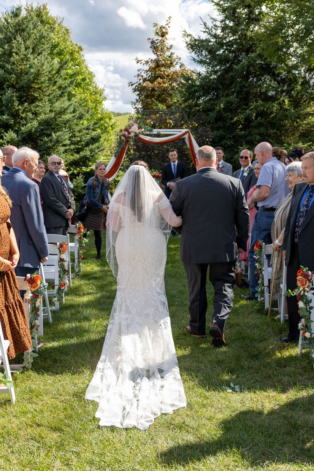 An outdoor wedding ceremony with guests seated on white chairs as a bride in a lace veil walks down the grassy aisle.