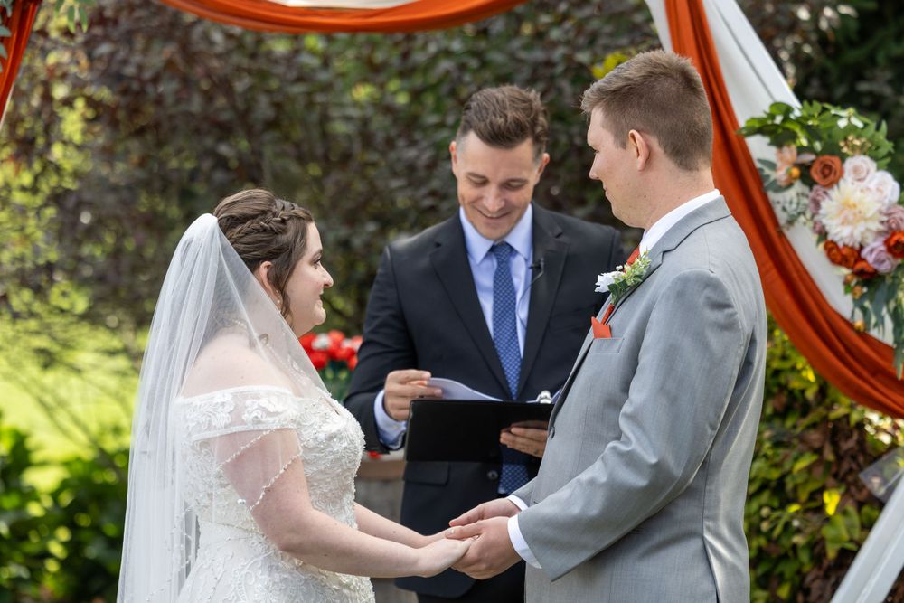 A wedding ceremony takes place outdoors under a red arch decorated with flowers as the couple holds hands before the officiant.