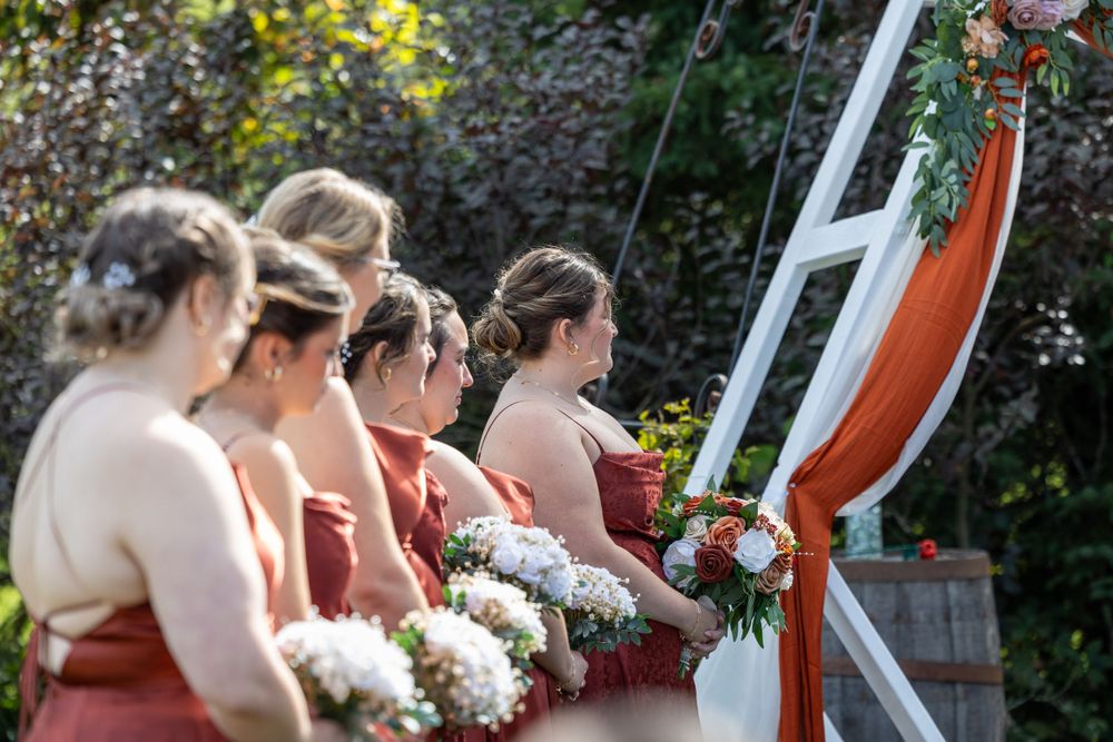 A row of bridesmaids in coral dresses hold white bouquets at an outdoor wedding ceremony.