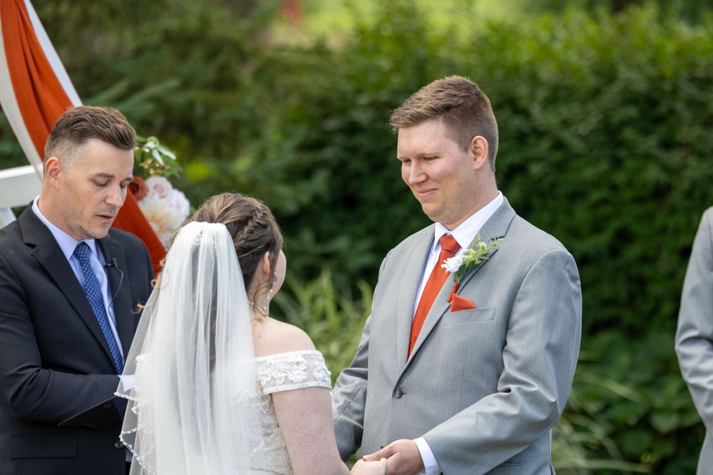 An outdoor wedding ceremony with attendees in formal attire against a green garden backdrop.