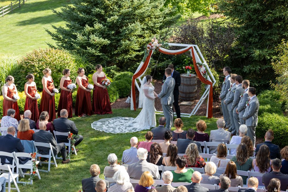 An outdoor wedding ceremony takes place under a wooden arch surrounded by evergreen trees and guests seated on white chairs.