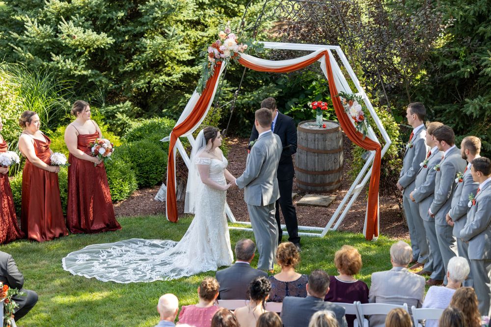 Outdoor garden wedding ceremony under a rustic arch with burgundy bridesmaids and gray groomsmen surrounding the couple.