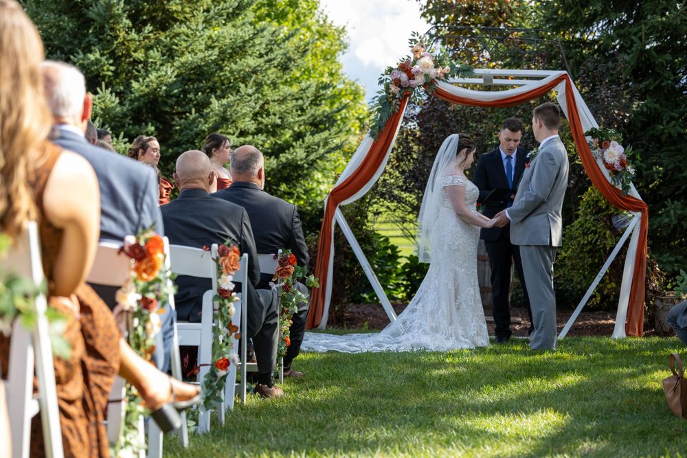 An outdoor wedding ceremony under a draped arch with guests seated on white chairs in a garden setting.