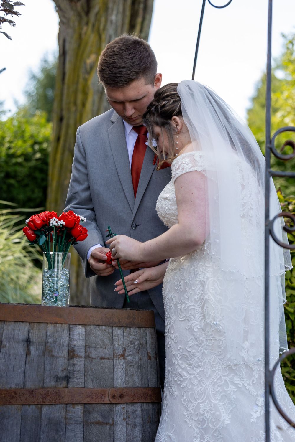 A tender moment at a wedding ceremony as a couple stands by a rustic wine barrel with red flowers in a vase.