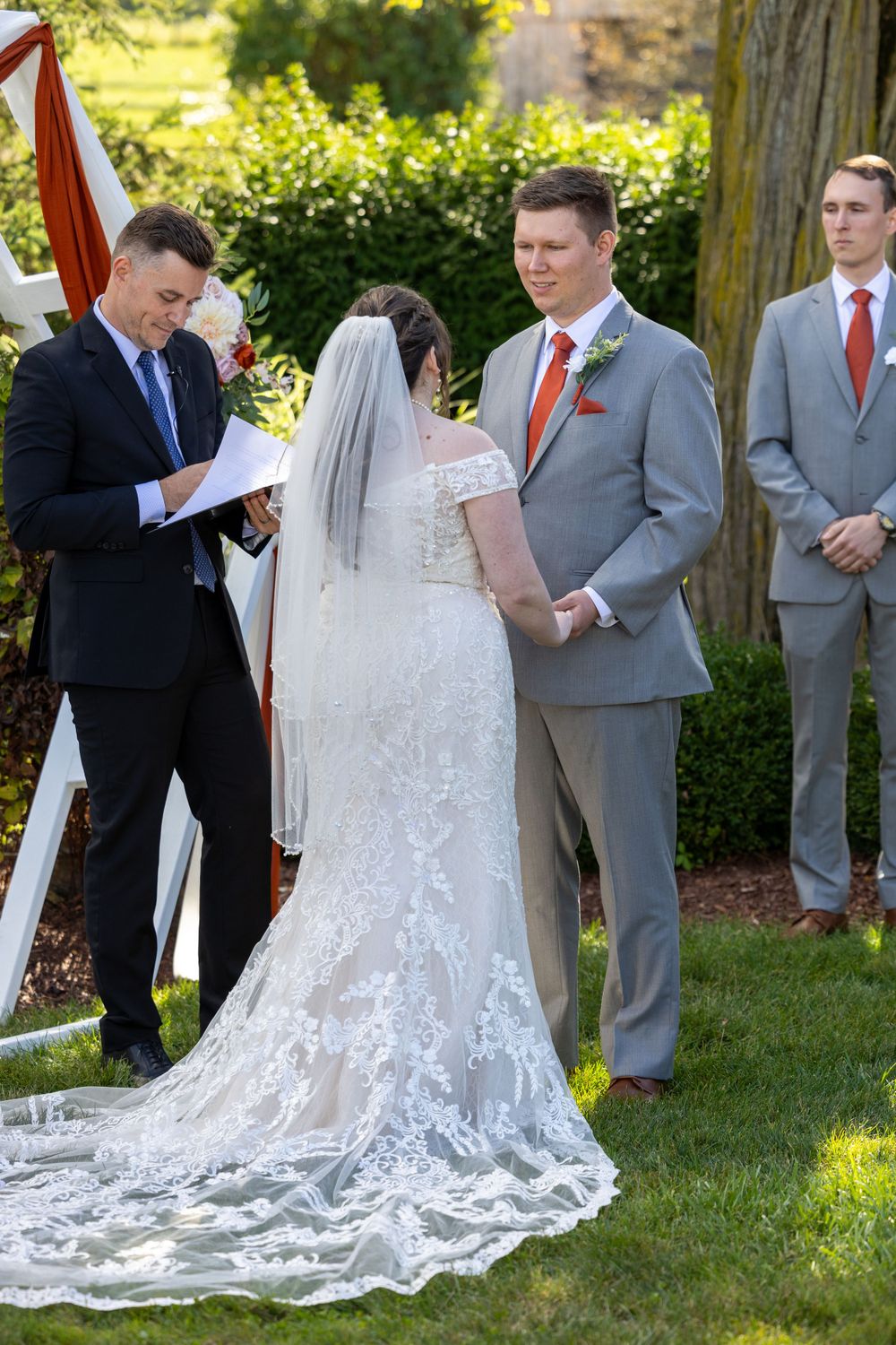 A wedding ceremony takes place in a garden setting with a stunning lace train trailing behind the bride's dress.