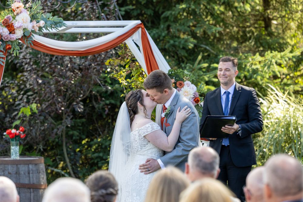 An outdoor wedding ceremony taking place under a decorative white and red draped arch in a garden setting.