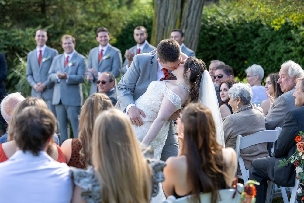 A romantic wedding ceremony kiss in front of seated guests with groomsmen in gray suits looking on.