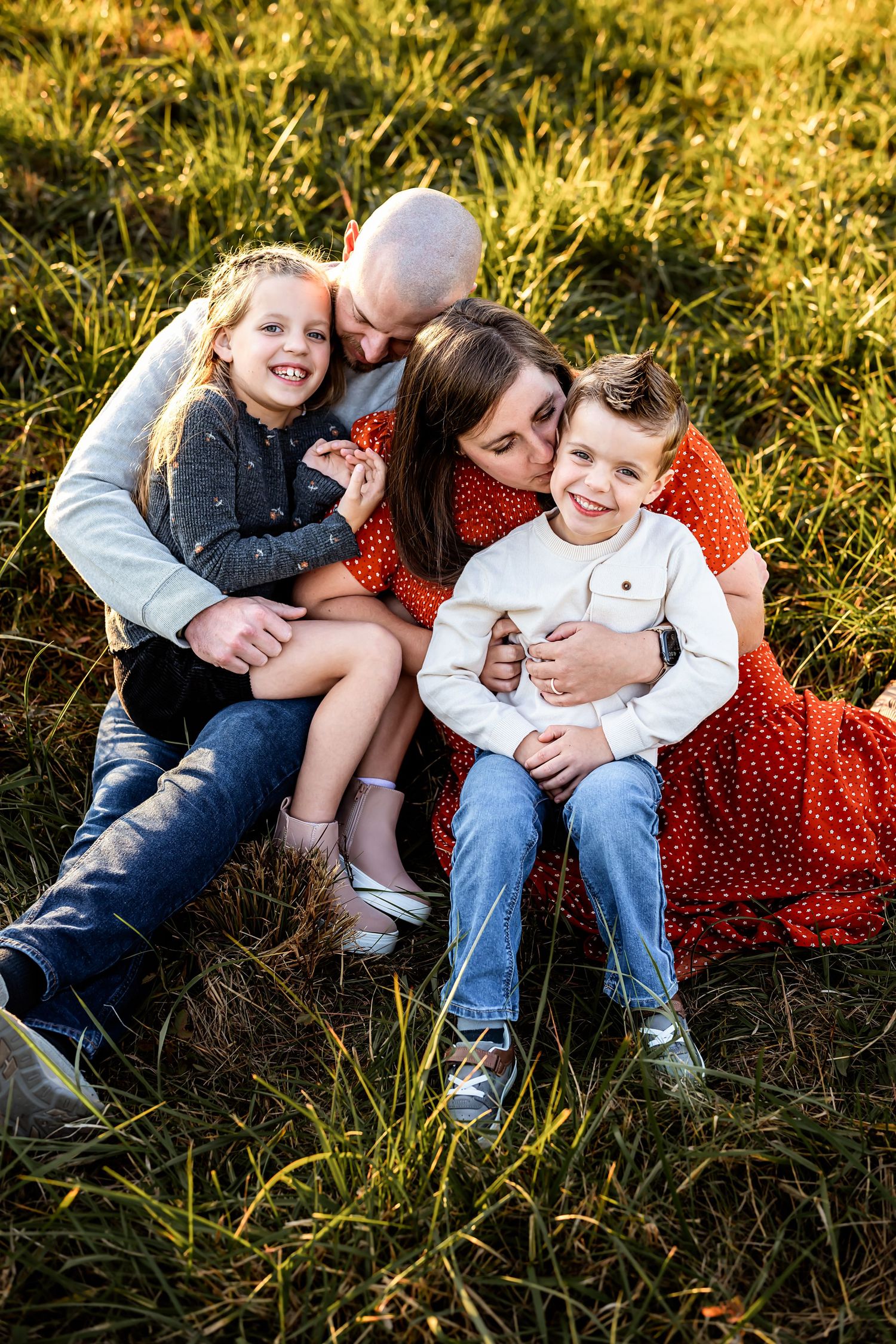 A joyful family laughs together while sitting in tall grassy field during golden hour.
