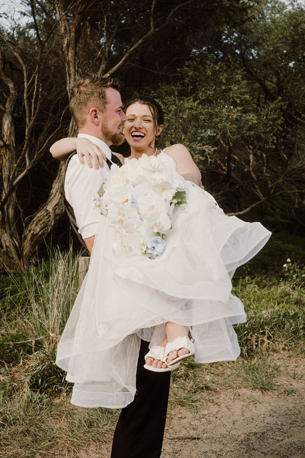Joyful groom lifting bride in white dress during outdoor portrait session.