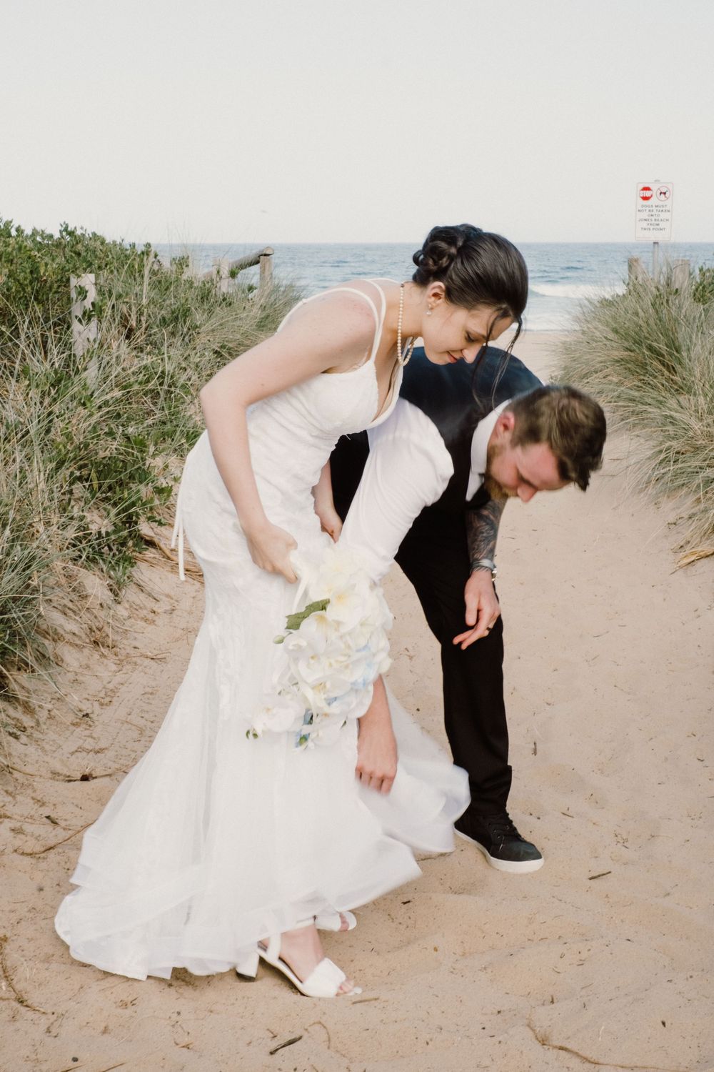 Newlyweds sharing playful moment on sandy beach path with white bouquet.