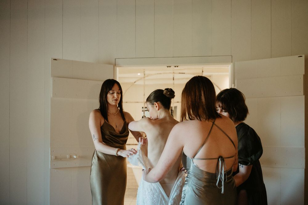 Wedding party helps bride get ready in elegant room with natural lighting.