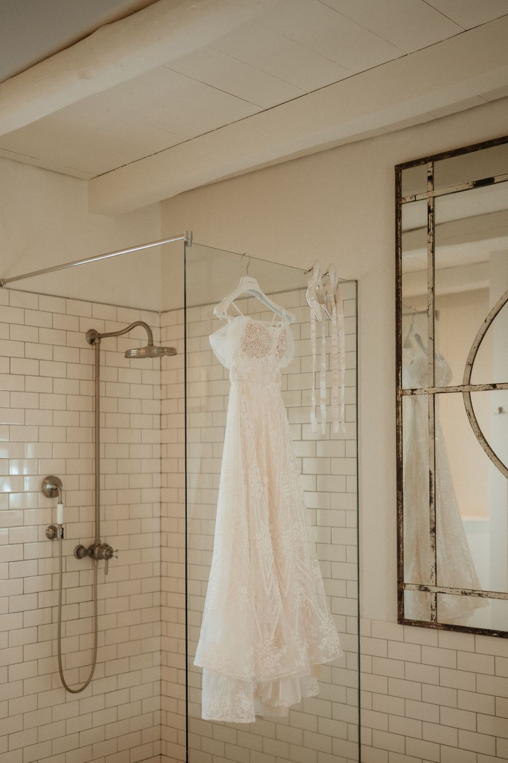 A delicate white lace wedding dress hanging in a modern bathroom with subway tile and glass shower.