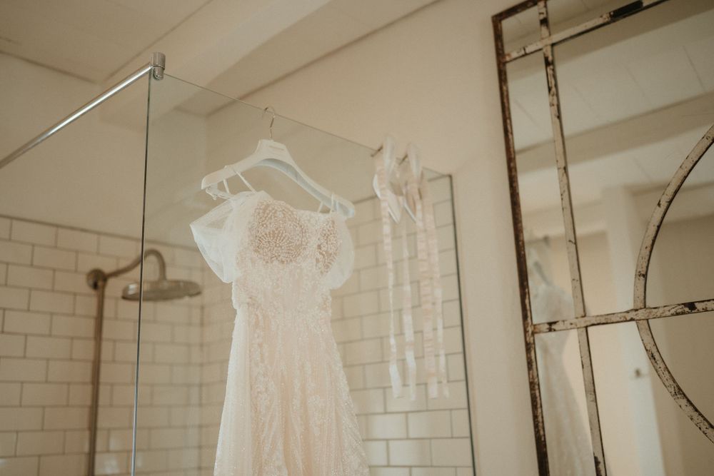 Delicate lace wedding dress hangs in a white-tiled bathroom.
