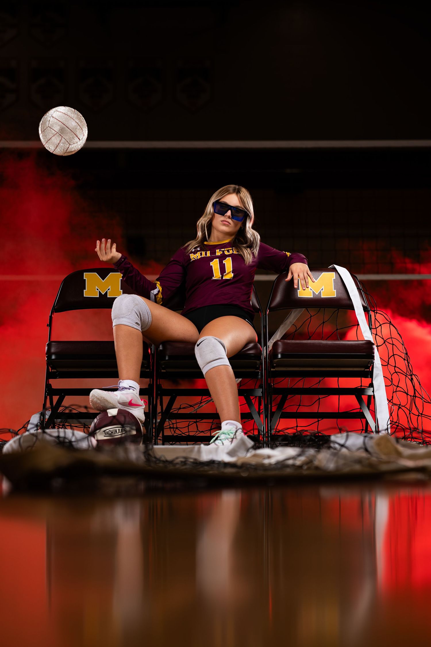 Softball catcher in protective gear poses with ball against dramatic white spotlights and smoke effect.